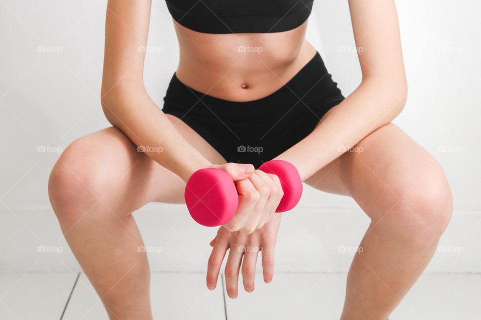 A young Caucasian girl in a half-crouch holds a pink dumbbell in her hands, doing an exercise against a white wall, side view close-up. The concept of a healthy lifestyle, sport and fitness