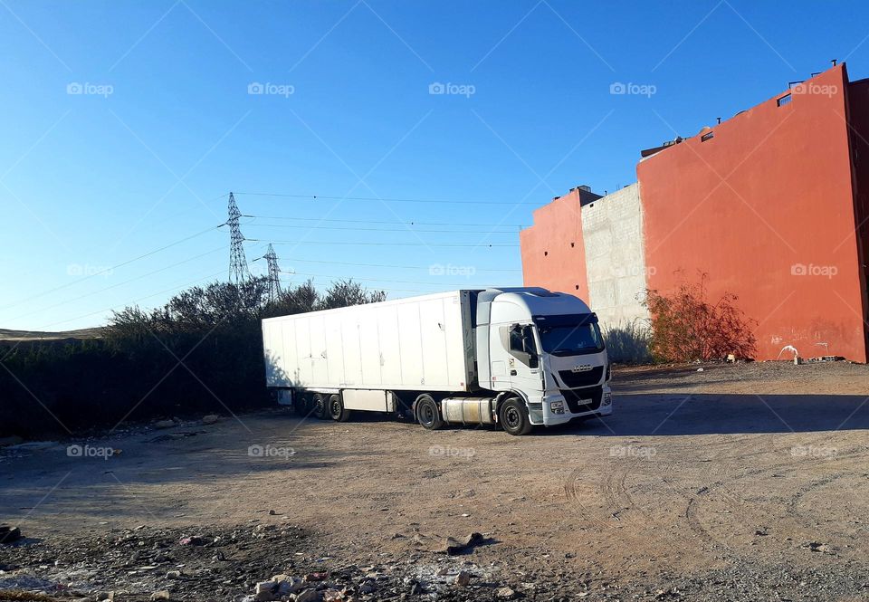 A white semi-truck is parked on a dusty lot near a large red building in Marrakech. The background features power lines, bushes, and a clear blue sky. The scene reflects an industrial or transport setting. January 1, 2025