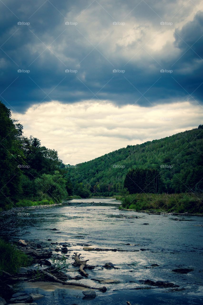 A dark and moody landscape photograph of the semois river in Belgium. the river flower through a forest and is in between mountains. the dark moody clouds set the scenic mood.