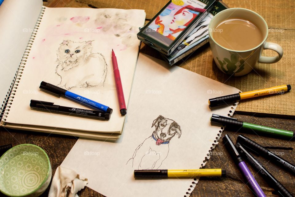 Favorite things: drawing cat and dog pet portraits at home with favorite Faber-Castell PITT Artist pens high angle view of sketches on sketch book paper on wood table with coffee, water for watercolor technique and blotter