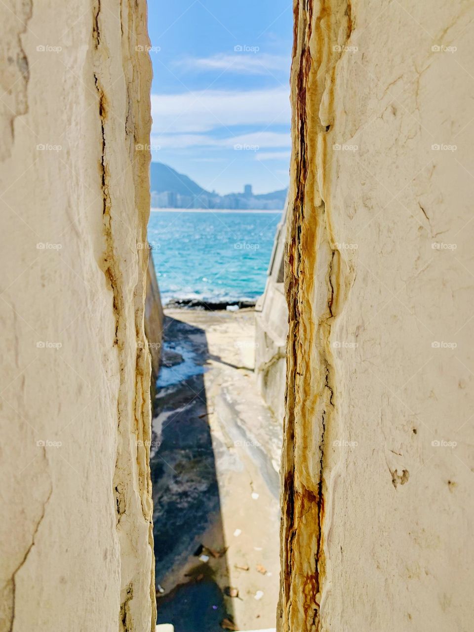 A picturesque view of the coast seen through the crevice in worn stone walls, highlighting the contrast between rough textures and the vibrant blue of the sea and sky in the background.