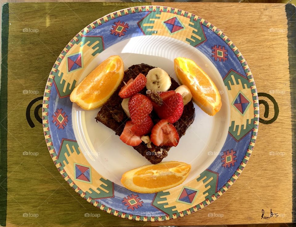 French toast with slice of strawberries, bananas on top. Walnuts, raisins and slice of oranges in a designer round plate with brown-green background.