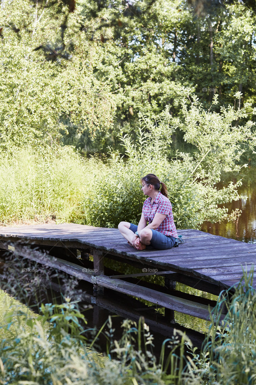 Woman sitting on a bridge over a lake, among the trees, close to nature, during summer vacations. Candid people, real moments, authentic situations