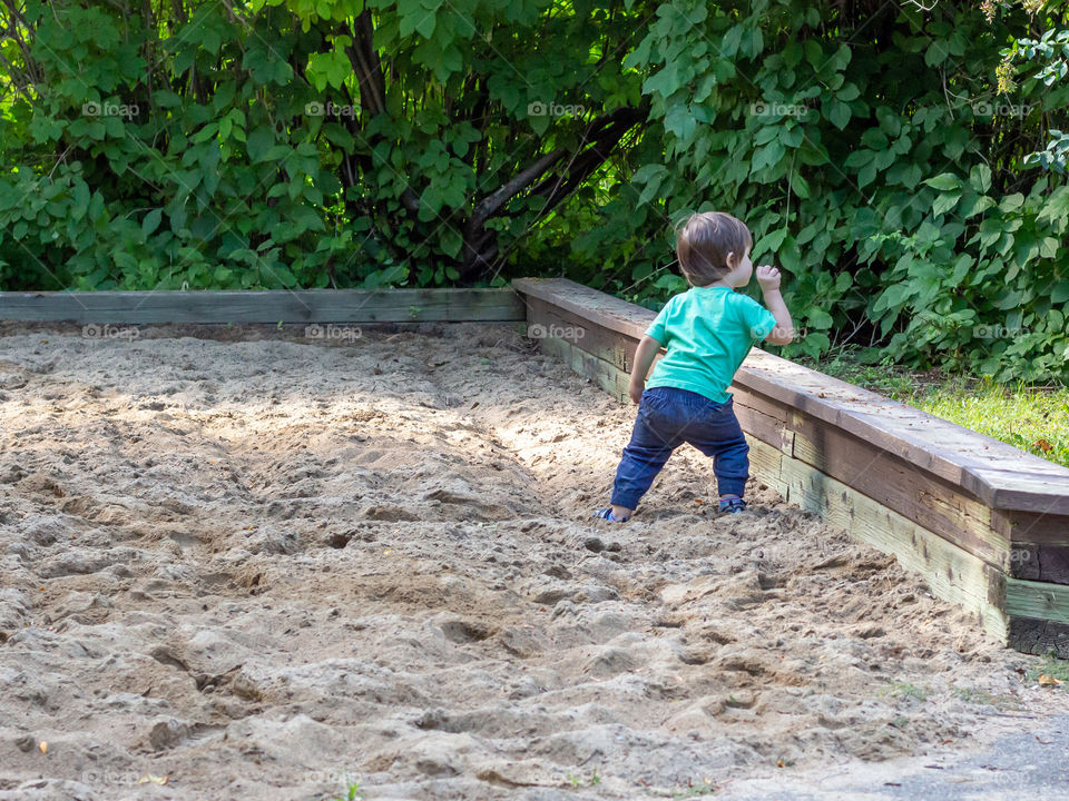 A little boy, about two years old, throws sand from a sandbox in a public park into the surrounding grass. The toddler faces away as he takes this defiant action, one he was just told not to take.