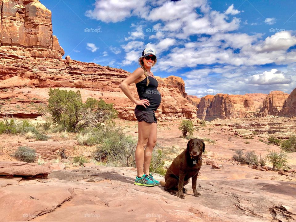 A pregnant woman and her dog stop to pose for a photo while hiking in the dessert on a beautiful spring day. 