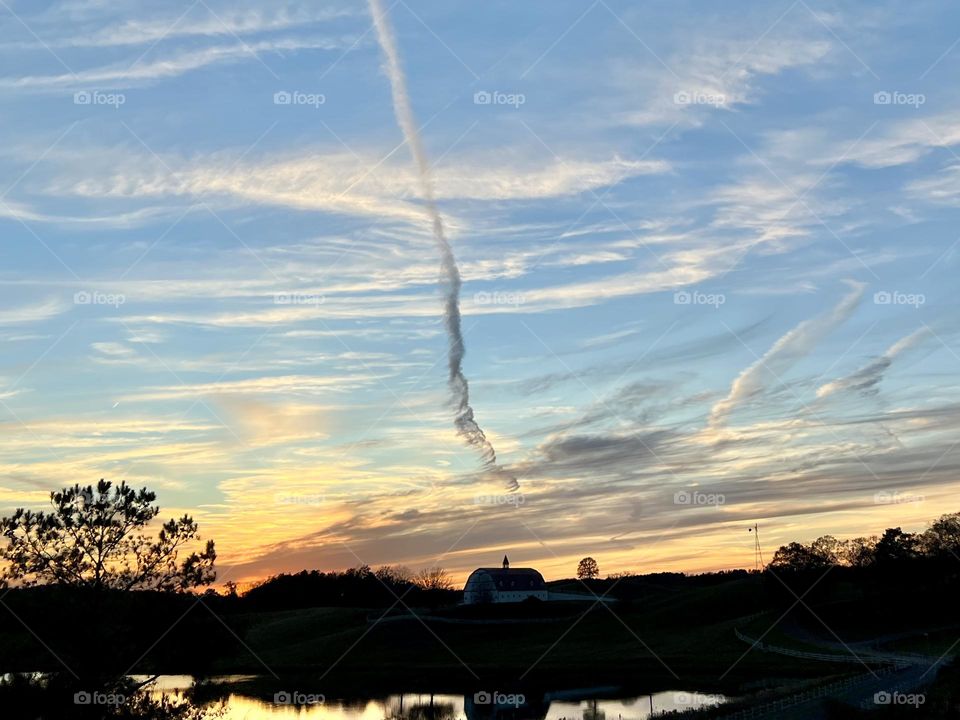 Streaking and swirling clouds create a dramatic sky at sunset. The view is silhouetted bay barn, windmills and trees, as well as reflected in a foreground lake