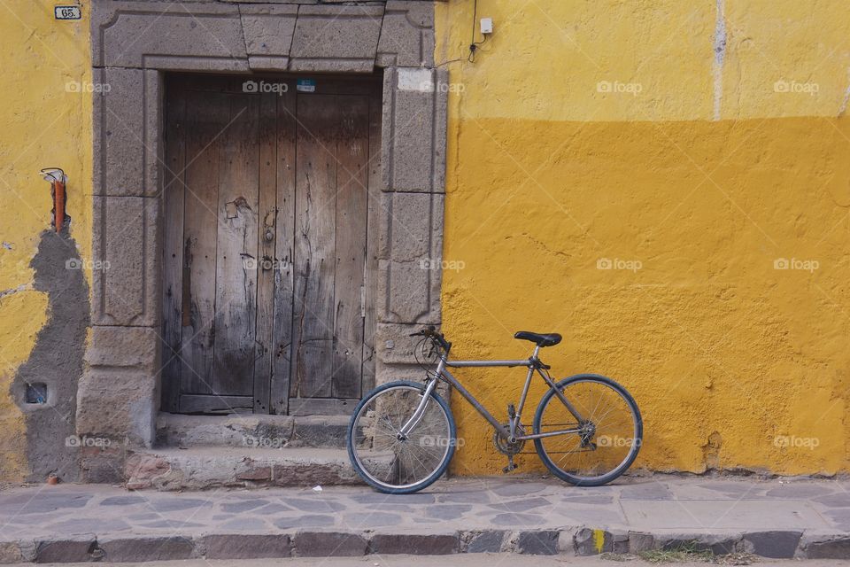 A bicycle is parked outside a  colorful yellow concrete wall surrounding a worn wooden doorway.