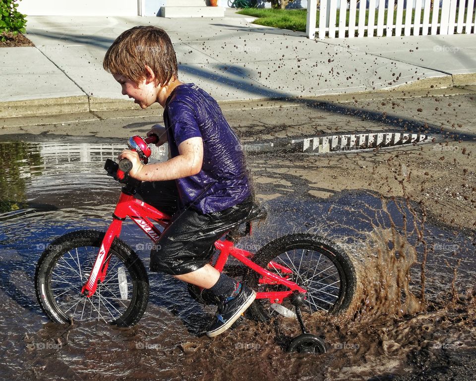 Getting Dirty In The Mud. Boy Riding Bike Through A Puddle During The Golden Hour
