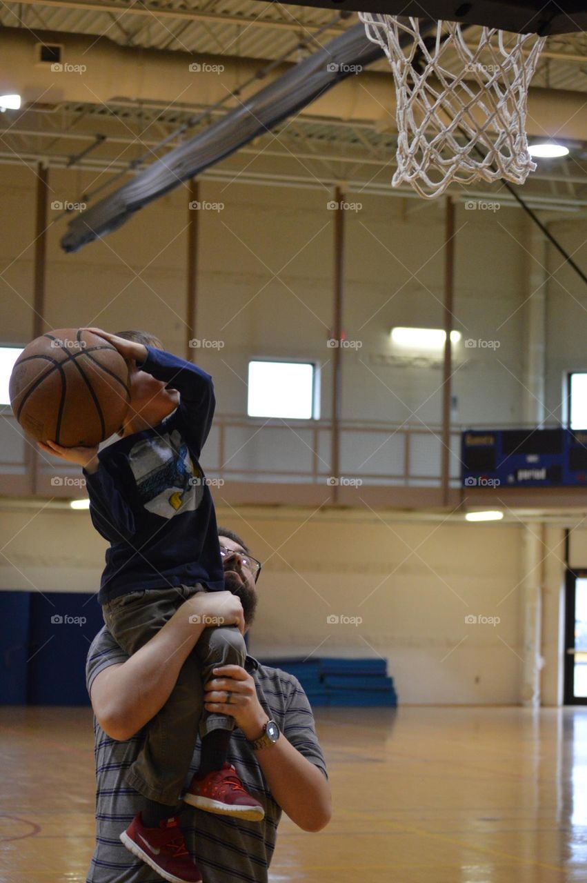 Young toddler boy on a basketball court shooting a basketball from his father’s shoulder 