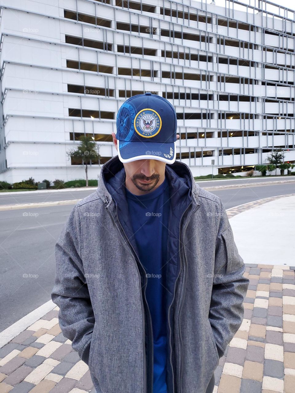 Demi saison. A man standing with his hands in his gray fall jacket pockets. He is wearing a blue sports shirt and a matching blue cap. A casual sporty fall season look in the city with a white parking garage building in the background.