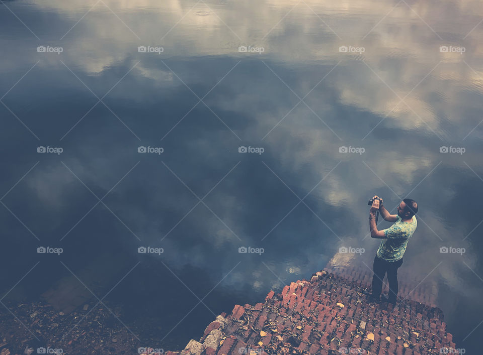 A somewhat abstract image that plays with perspective. The shot is downwards looking at a man standing at the bottom of some steps. He is taking a photo of the river which dominates the rest of the image.The clouds reflecting in the river confuses us