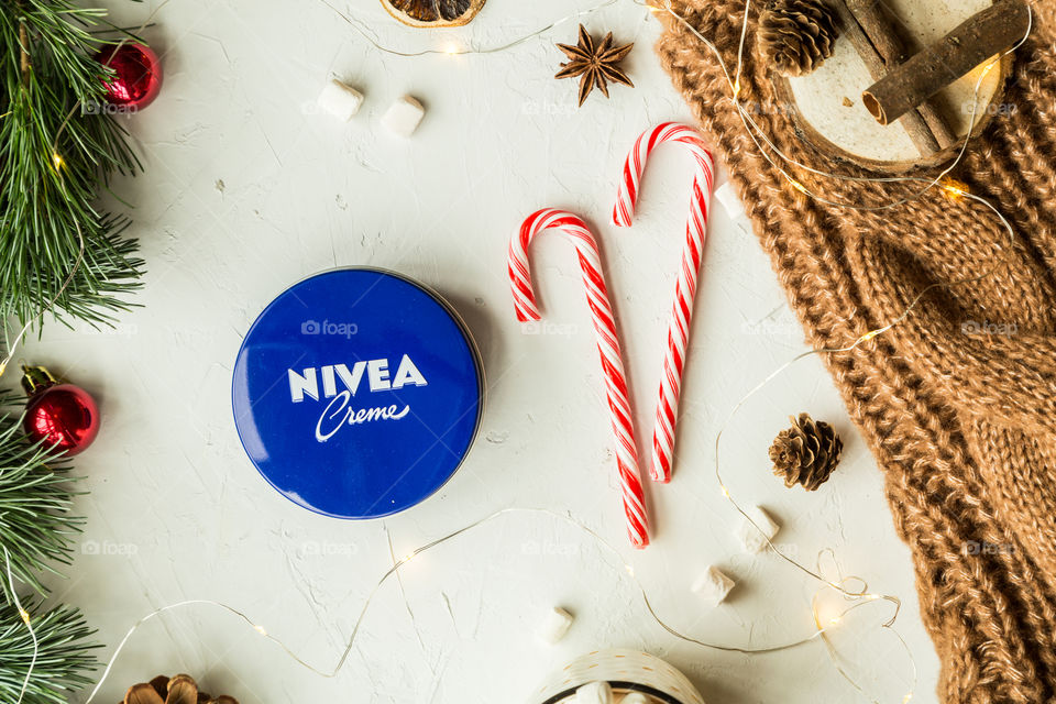 Christmas time with Nivea cream. Flatlay, top view, cup with marshmallows, dry orange, cardamom, cinnamon, pine cones, sweety, tree, garland, knitted sweater brown color, red balls . Top view 