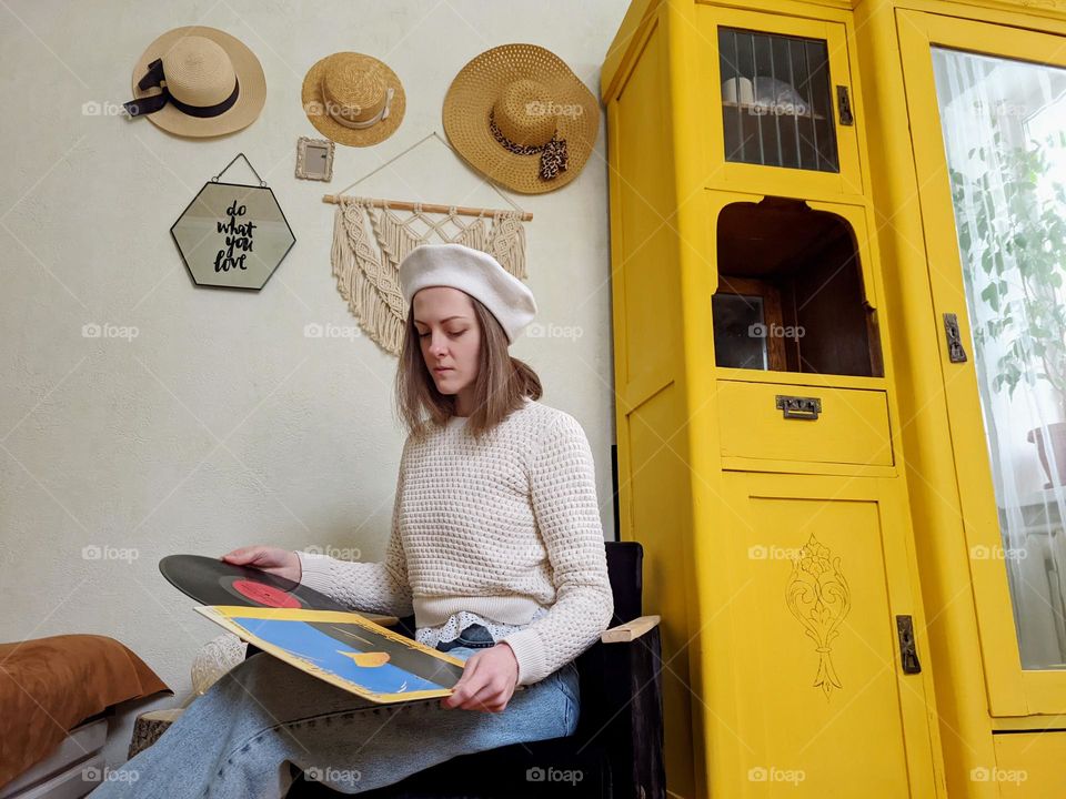 Portrait of beautiful young girl,  lady in beret sitting at cozy home in armchair near yellow vintage cabinet.  Vinyl music