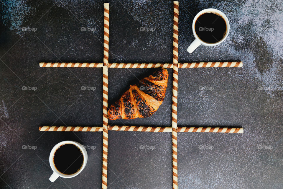 Business working morning with cup of hot coffee, sweet croissants on black background close up. Game of noughts and crosses. Top view, copy space, flat lay, mockup.