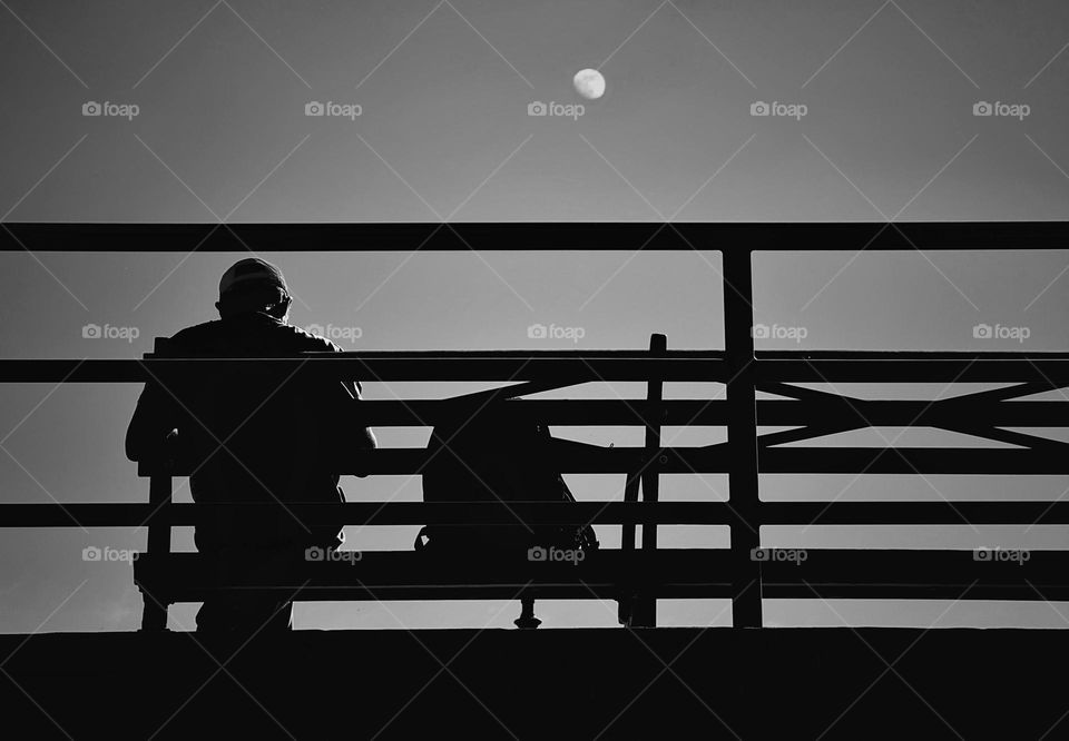 Black and white silhouette of a man sitting on a bench with the moon above in an afternoon sky