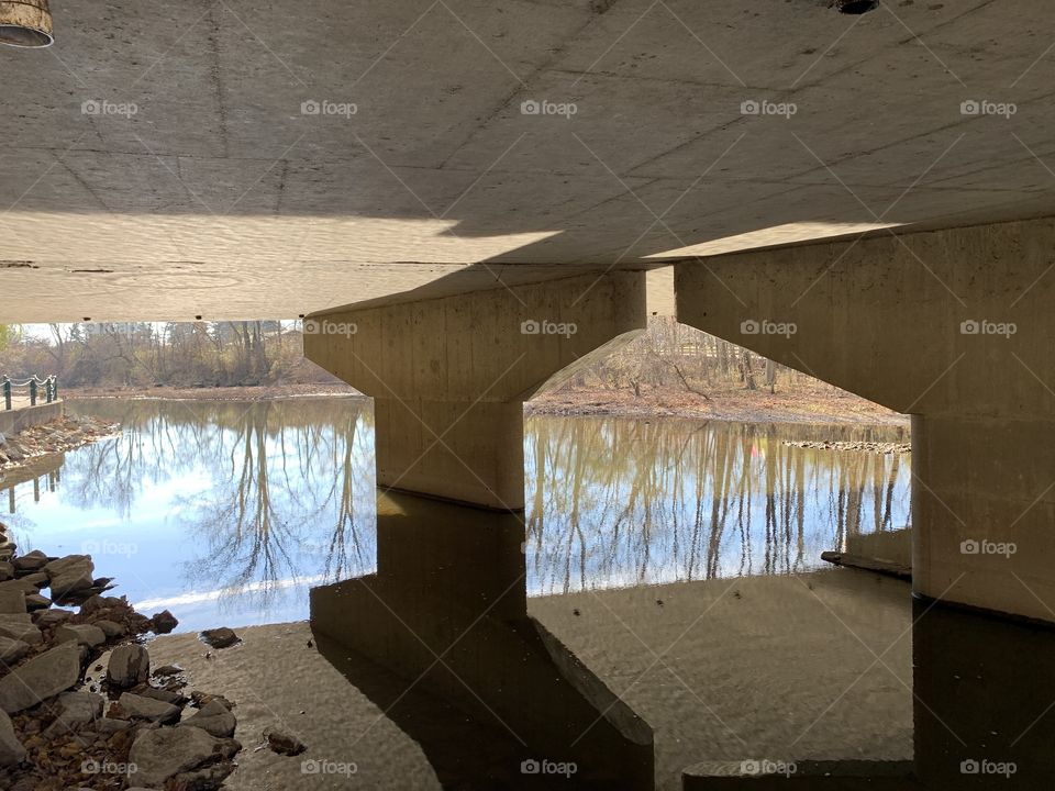 A bridge over tame waters. These waters are just about clear enough to see to the bottom. I can see my reflection clearly, as well as the rocks beneath. 