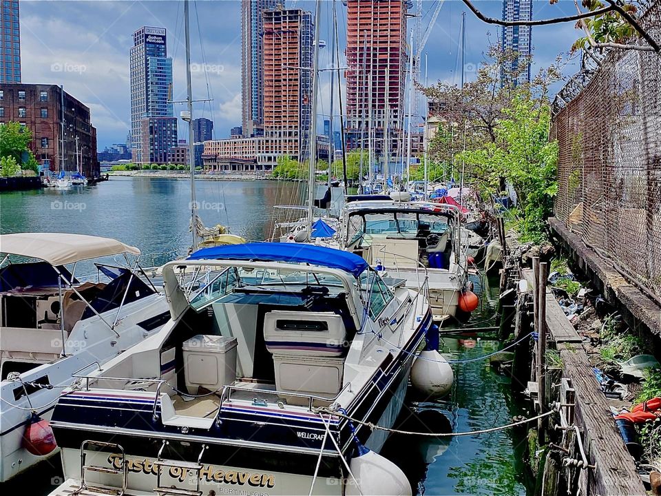 Boats are lined up side by side along the wall at „Newtown Creek“ by the „Pulaski Bridge“. Lush green trees accentuate this scenario and make it appear even more idyllic. 2023. Hypnotic Productions