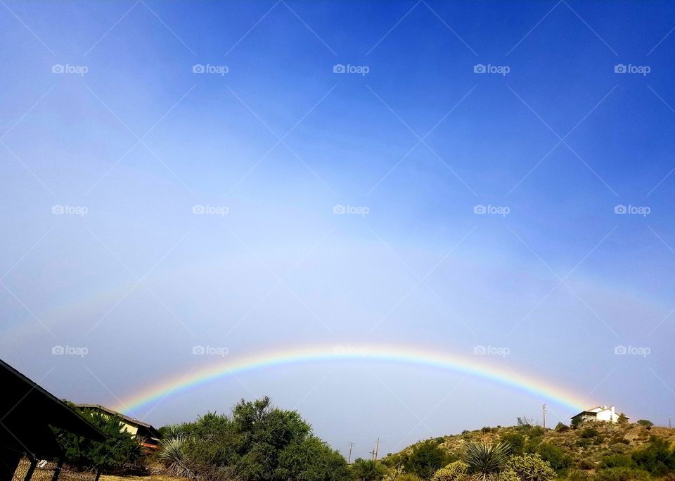 Double rainbow stretched between 2 houses. Brilliant blue sky and landscape frame the rainbow.