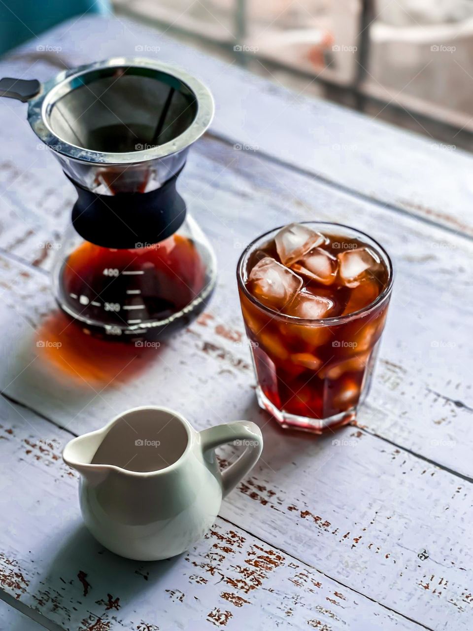 Iced Americano in a cup next to coffee dripper and a small milk jug on white wooden table
