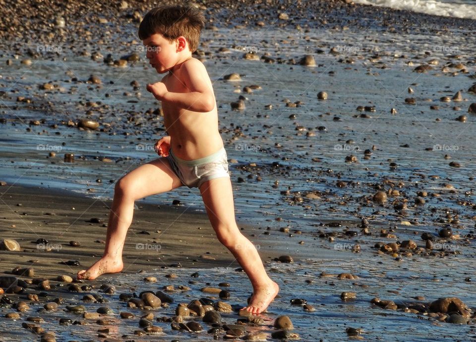 Little Boy Running Barefoot On A Pebble Beach. Running From The Sea