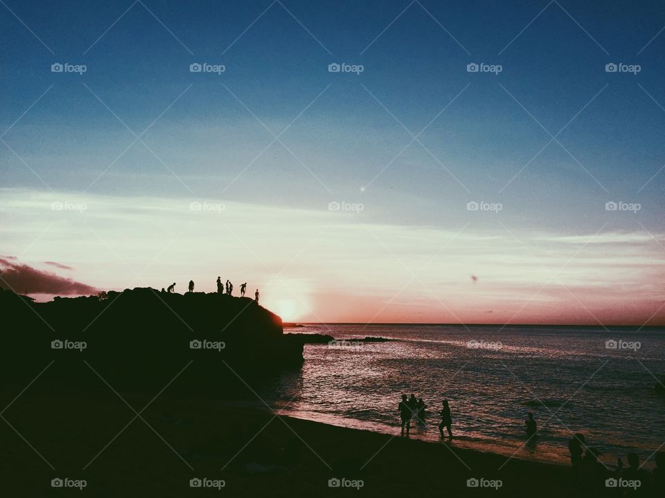 People cliff jump on the north shore of Oahu, Hawaii during a sunset over the ocean.