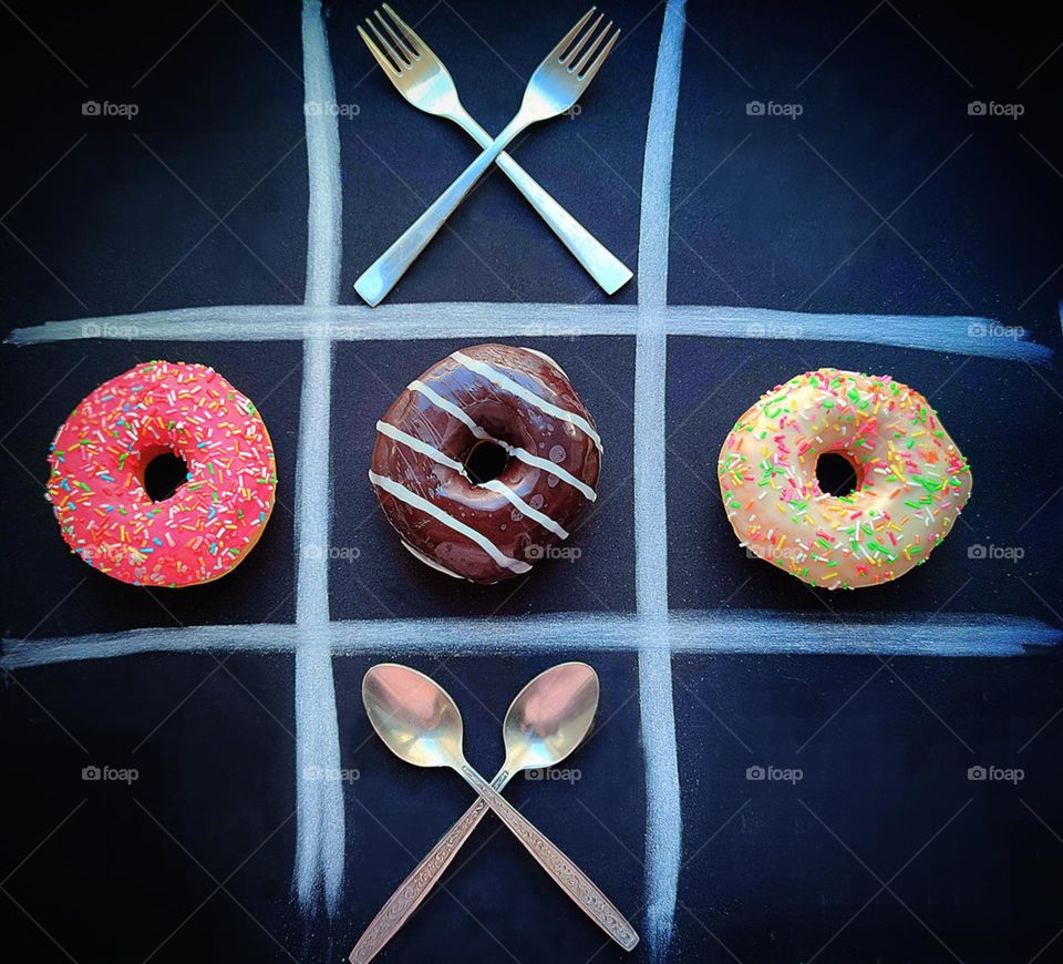 Food. Tic-tac-toe. The black background is lined with white. Diagonally in three cells are donuts of different colors (zeros). In the upper cell are two crossed metal forks (cross). In the bottom cell are two crossed metal spoons (cross)