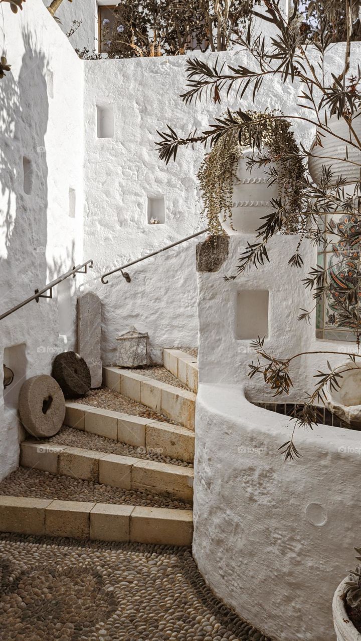 Beautiful outdoor interior of an ancient Greek street in Lindos city: paved pavement with small stones, round house and spiral steps leading to the right with olive branches and flowers on the island of Rhodes, close-up side view.