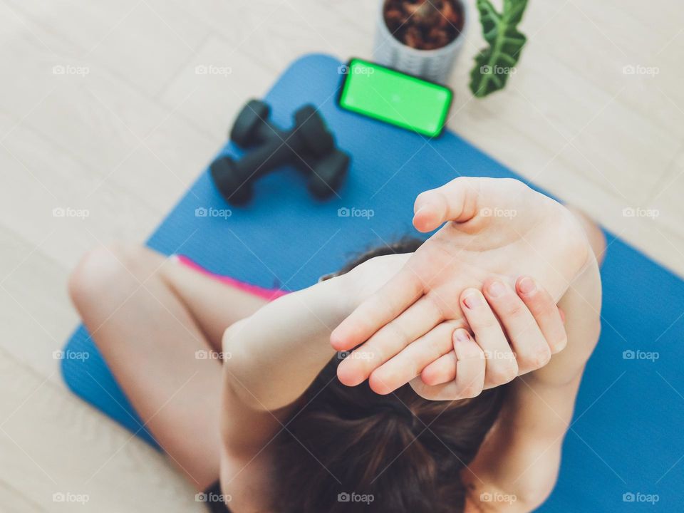 Young caucasian girl doing exercise sitting on a mat on the floor with a green screen phone, dumbbells and a flower in a pot, top view close-up with selective focus.