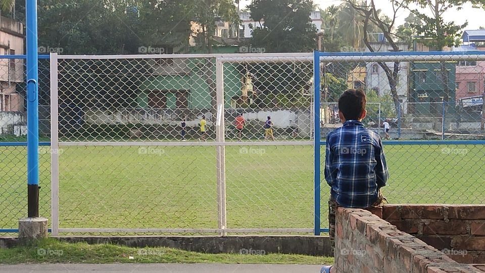 One boy is sitting and watching the green field alone. The playground is fenced with a net.