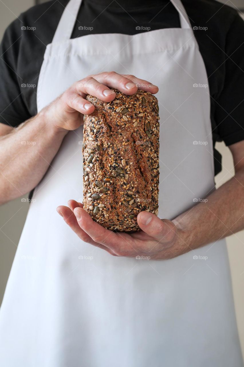 Close-up at bakers hands holding a multi-seeded loaf of whole grain bread in front of him.