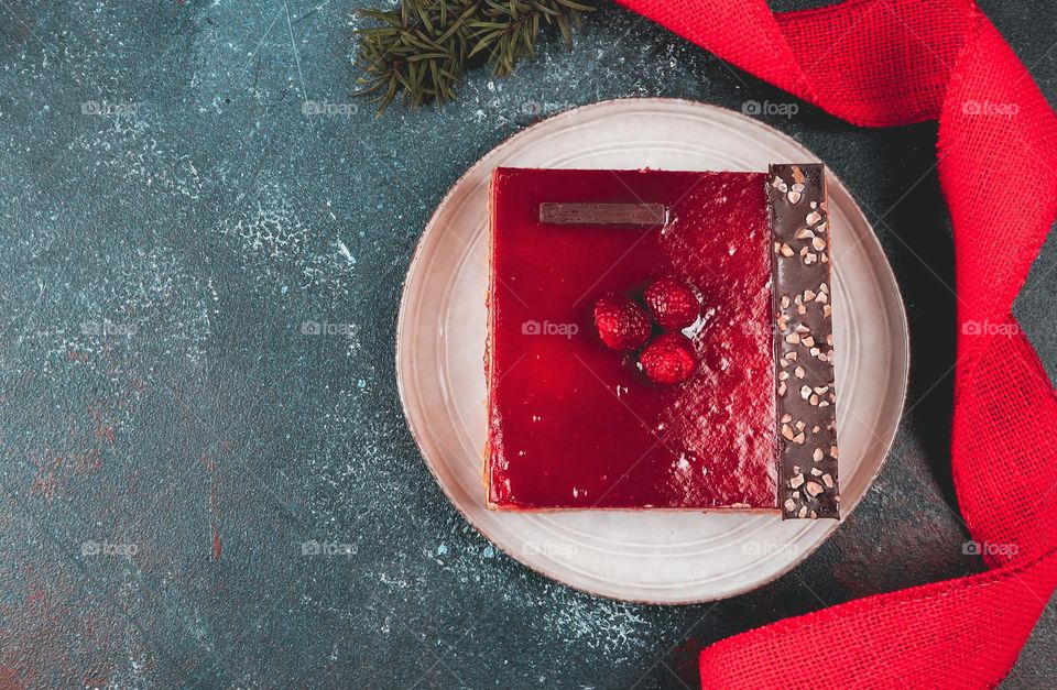 One square raspberry cake on a round plate with a red napkin on the right on a dark cement floor with copy space on the left, flat lay close-up. Holiday food concept.