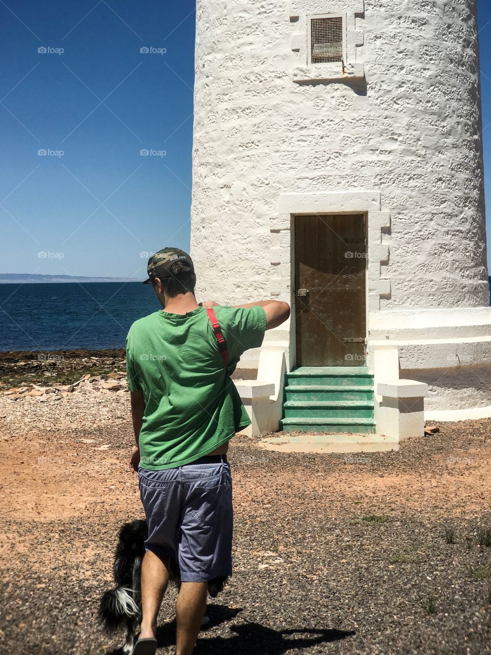 Young man in tee shirt and shorts walking dog toward old white stone lighthouse with green stairs and brass door