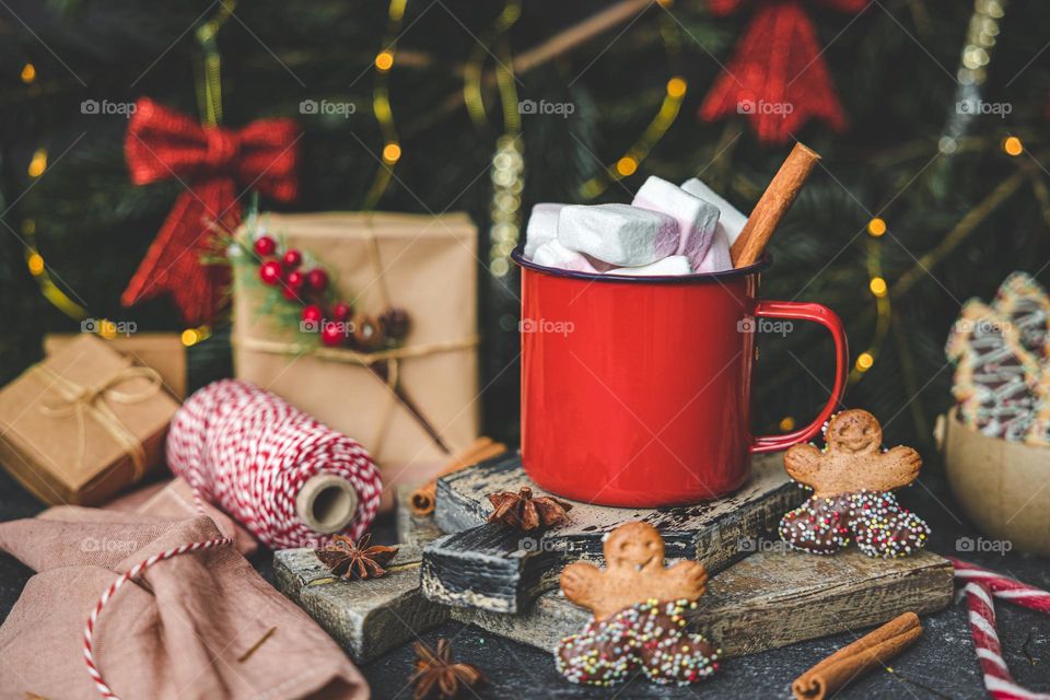 One red mug of hot traditional cocoa drink with marshmallows, cinnamon, anise, gingerbread man cookies and gift boxes with a spool of thread on a dark cement table with a blurred background of spruce branches, a bow decoration with a burning garland