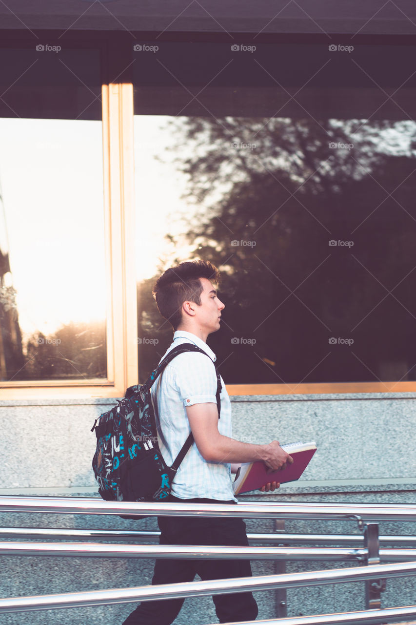 Student holding a notebook and carrying a backpack on his back walking at the front of university building. Young boy wearing blue shirt and dark jeans