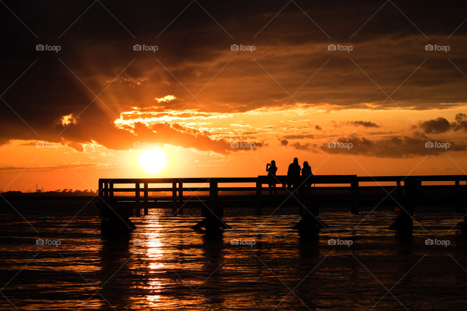 Friends enjoying a beautiful golden sunset on the water. Silhouetted figures standing on a pier under a vibrant orange sky