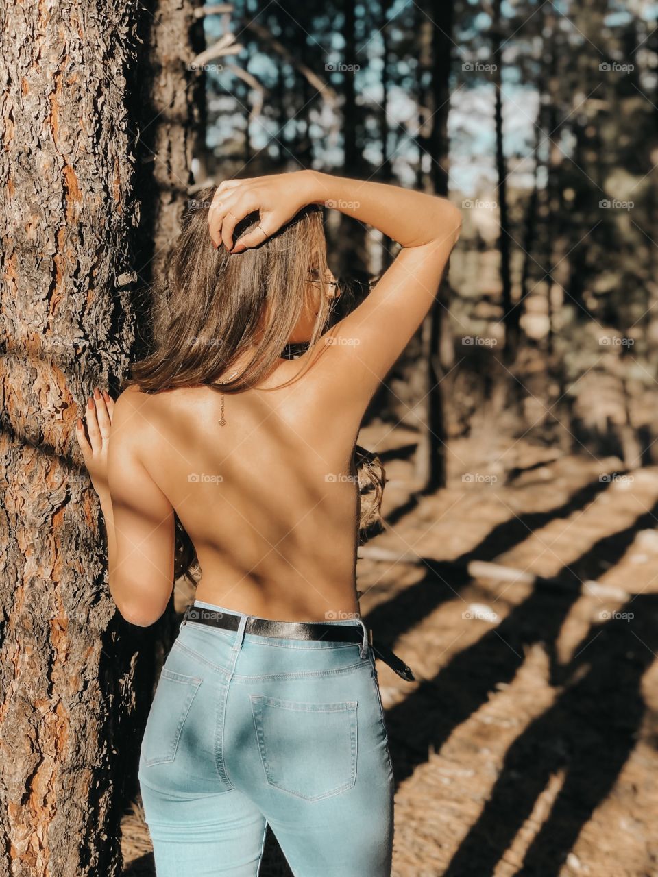 Young tan girl posing topless in the sunny shadows of the trees.