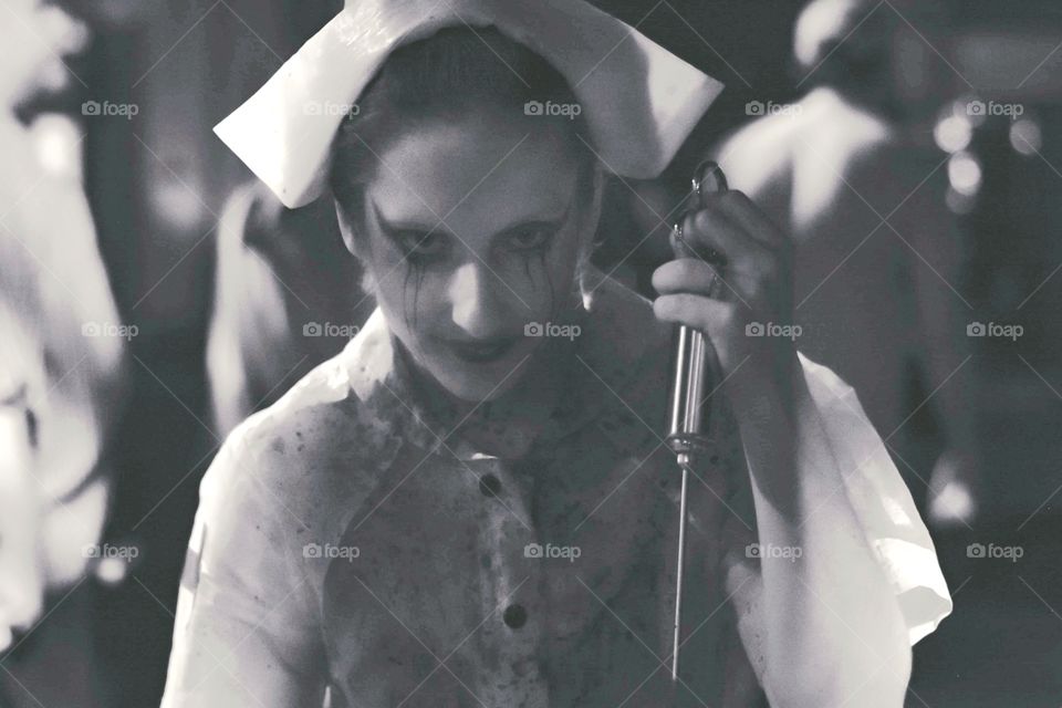 creepy black and white image of a nurse with blood stained uniform holding up a very large needle and looking into the camera