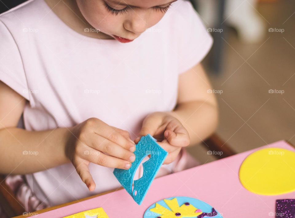 One little beautiful caucasian brunette girl in a pink t-shirt peels off felt stickers for an Easter craft, sitting at a children's table in her room, close-up side view. Creative kids concept.