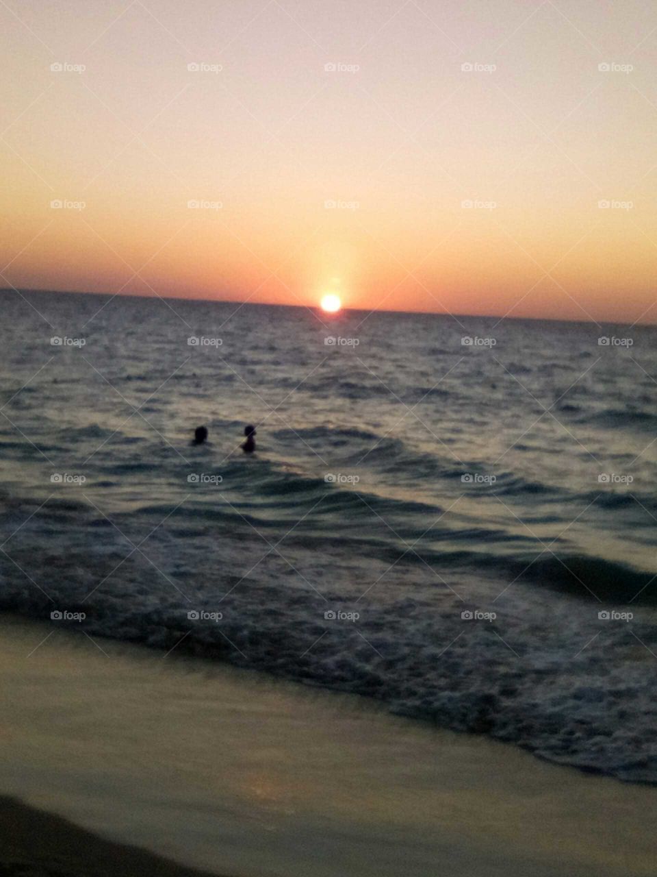 this picture is one of a kind  see how the sun sets behind the ocean the water an wave and the two people in water having fun .