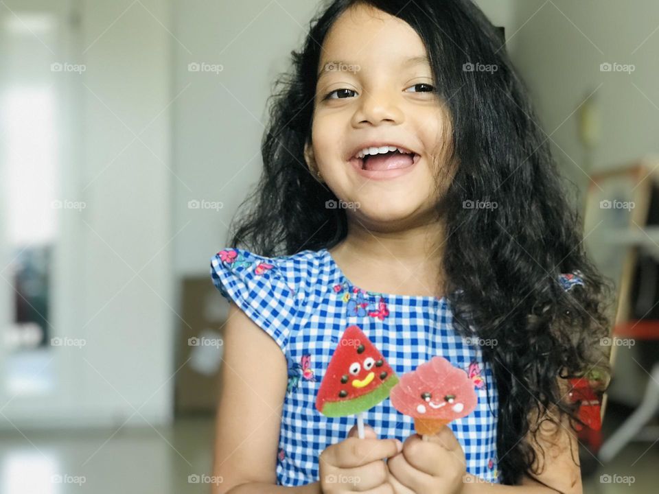 Four years old girl having fun by eating candy lollipops in summertime, she’s wearing blue colour frock and she have watermelon and ice cream candy lollipops in her hand,and she had a beautiful smile on her face.