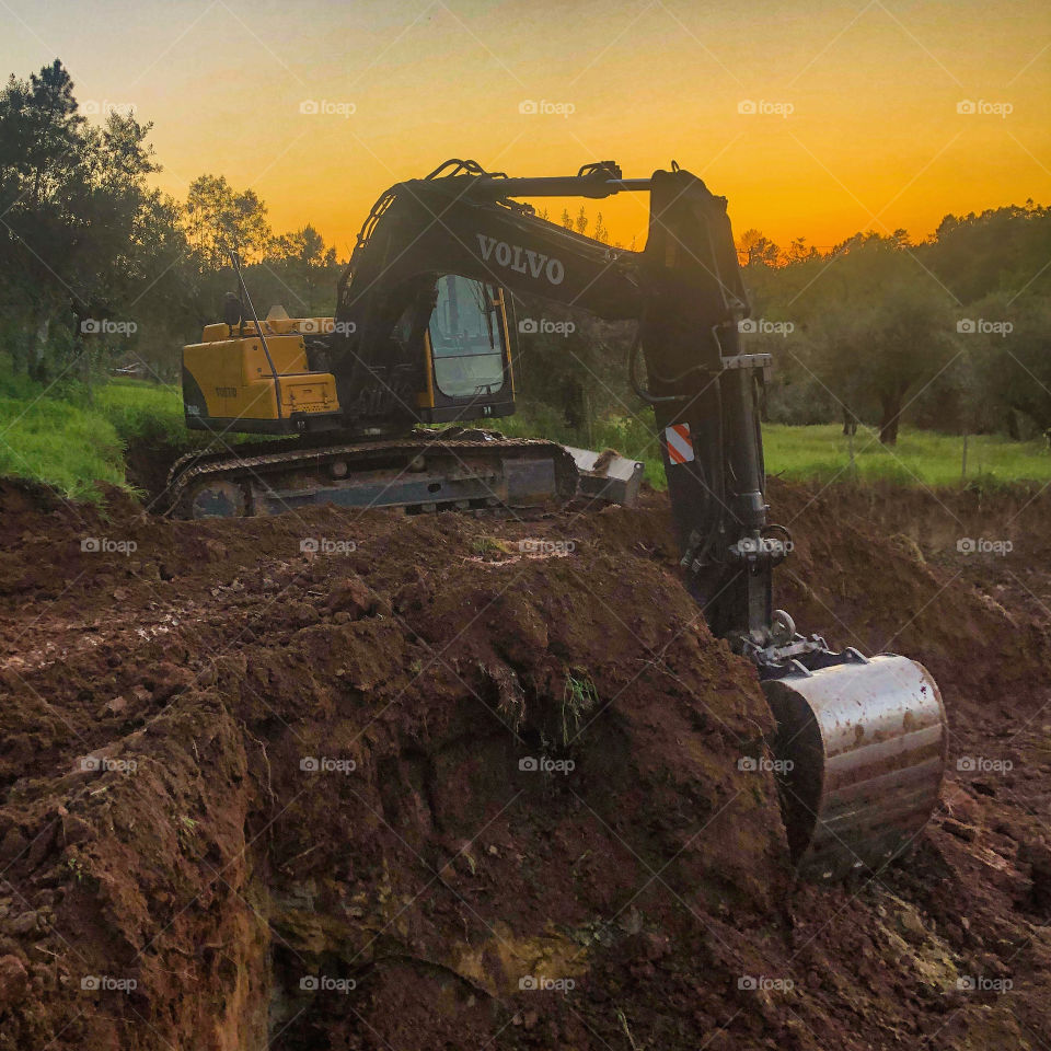 A metal digging machine sits at rest in the countryside at sunset