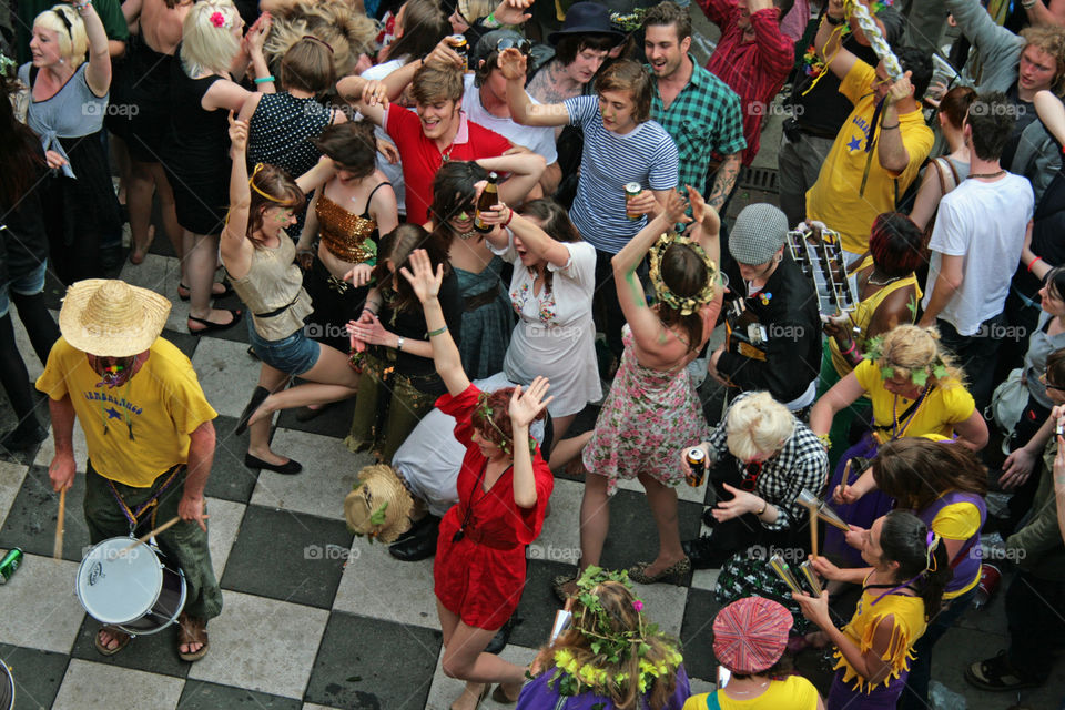 We look downwards to a group of young people who dance and celebrate on an outdoor checkered chessboard, they are dressed colourfully with flowers in their hair and a samba band plays - Hastings, U.K.