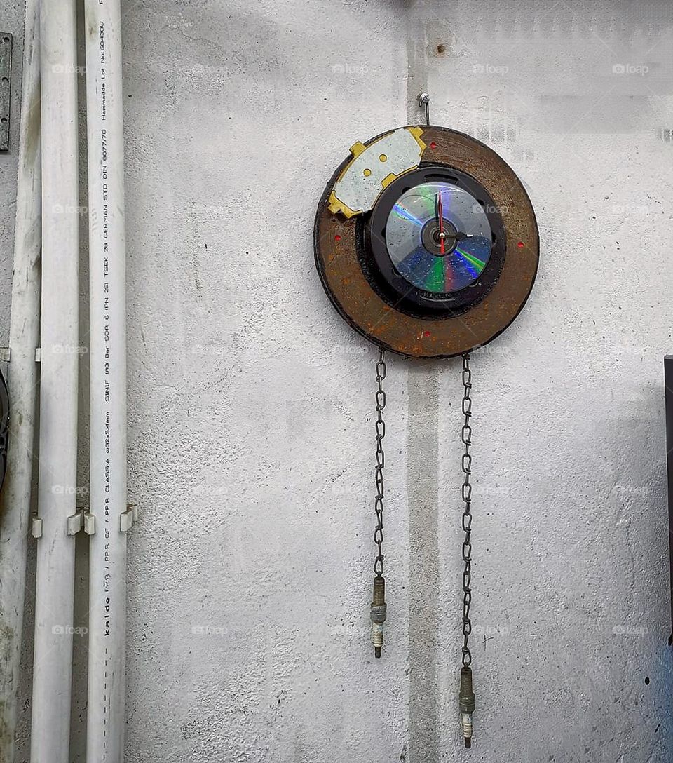 A clock is hanging on a white wall, which is assembled from a brake disc, a brake shoe, a DVD disc and spark plugs. Wall in a car workshop