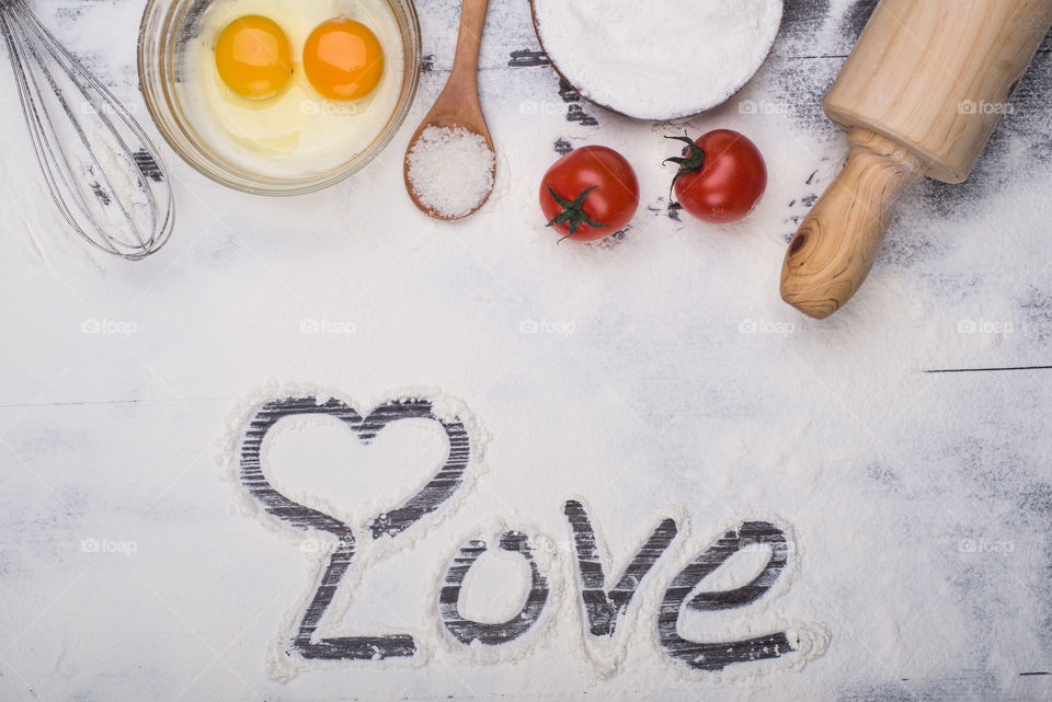Cooking needs a lot of love! Top view of table full of flour, along eith eggs, sugar, tomatoes and a molder