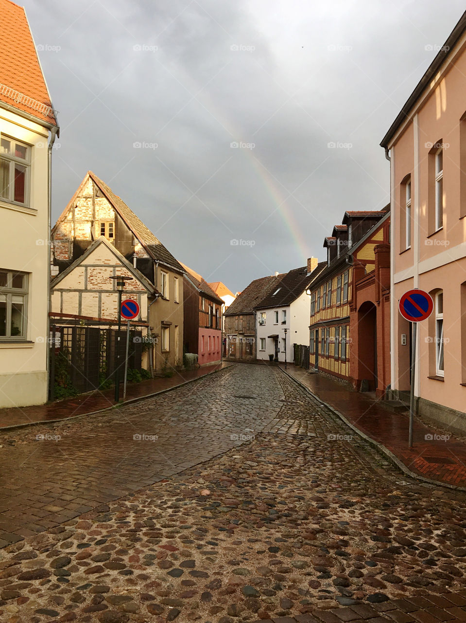 An empty street of a European city with old houses and paving stones.  In the background there is a rainbow in the sky.