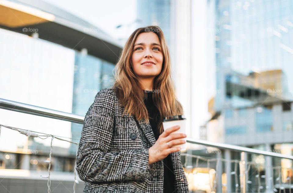 Young smiling woman in coat with paper coffee cup in evening city street