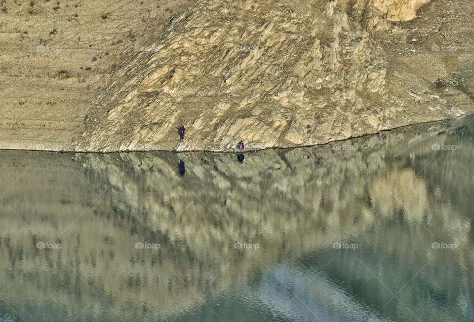 photo taken from a bird's eye view of two fishermen fishing near a mountain lake against the backdrop of a huge stone cliff. mirror reflection of the mountain gives the unreality of what is happening in the frame