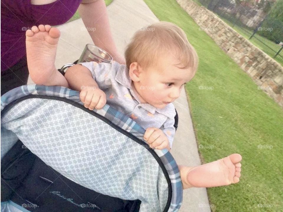 A curious baby sits whimsically on top of a stroller on an outdoor pathway, surrounded by green grass and a stone wall. Whee! Woohoo! 