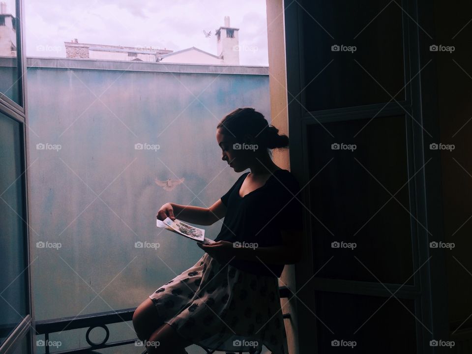 This photo is a gentle silhouette of a girl sitting in a window. This photo was taken in Paris, France. The gorgeous mural in the background contrasts with the dark inside of the room.