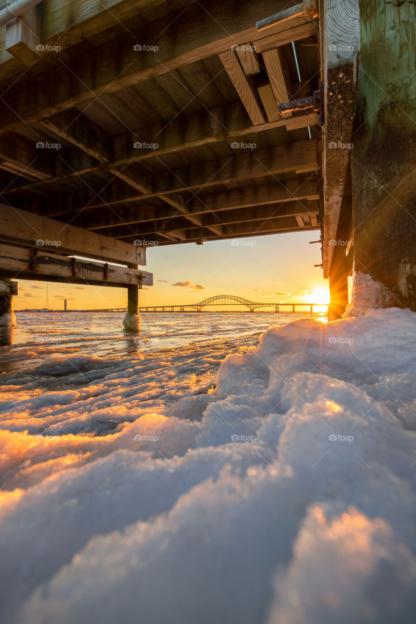 Snow and ice under a wooden pier as the sunsets in the distance, casting beautiful golden light on the winter scene. 